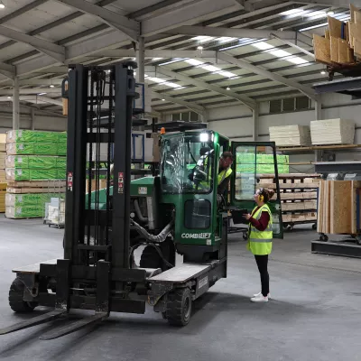 A person in a high vis vest with ear muffs show a tablet screen to a person driving a forklift.