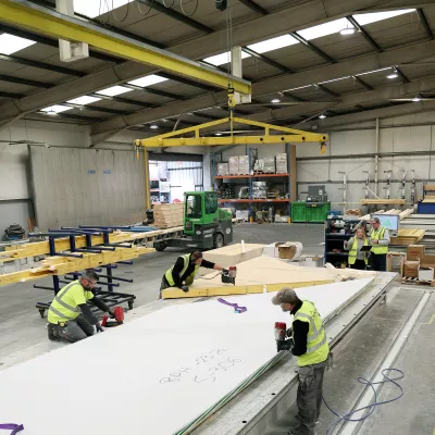 Three people in high vis jackets use power tools to cut wooden frames while two people in high vis jackets with a tablet observe.