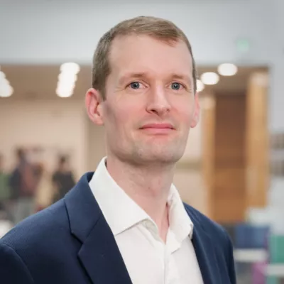 Headshot of a person in a suit, standing indoors with a blurred background.
