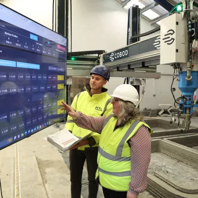 Two people in high-vis vests reviewing data on a large screen, with a 3D printer visible in the background.