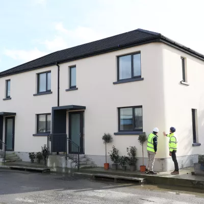 Two people in high vis jackets and hard hats inspect modern white semi-detached houses.