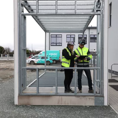A male and a female read from a tablet inside a prefabricated steel frame set in a car park.
