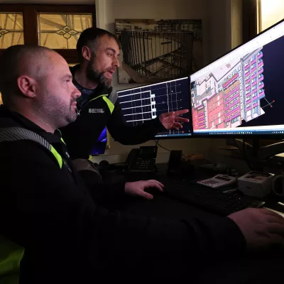 A male points out something in a digital building design on a computer screen to another male.