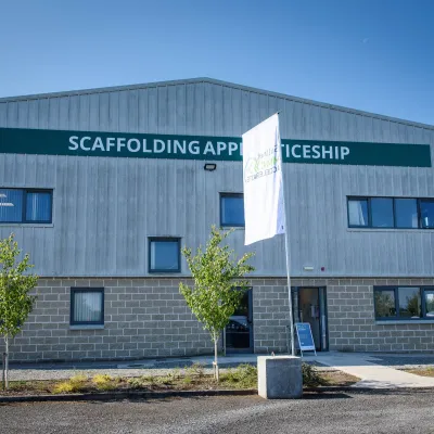 Grey building with the sign 'Scaffolding Apprenticeship' and two white flags showing the Skillnet MMС Accelerate logo.
