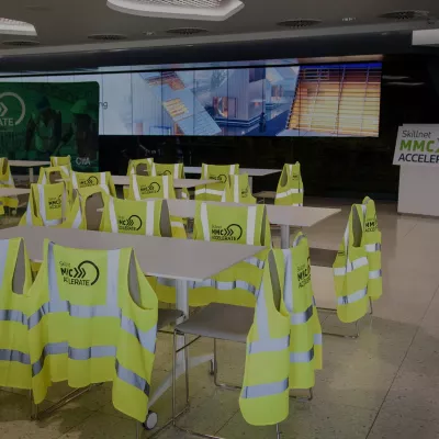 High vis vests with the Skillnet MMC Accelerate logo hang off the back of white chairs around white tables in an event space with a screen in the background.