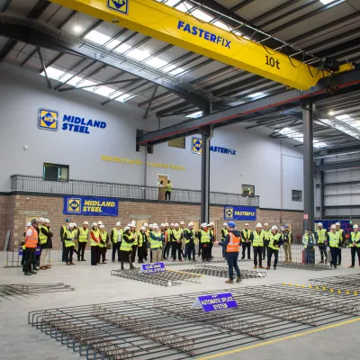 A large group of people in high-vis jackets and white hard hats inspect a a stack of welded steel frames.