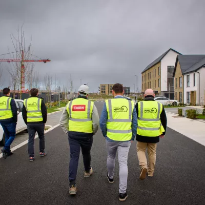A group of five people in high vis vests walk past a white car through a modern housing development.