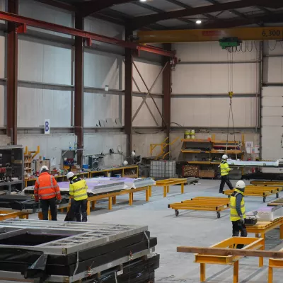 Several people in high vis vests work at machinery stations in a manufacturing facility.