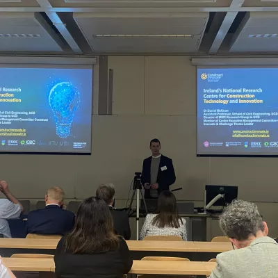Person presenting at a conference in a lecture hall, with slides projected on two screens at the front of the room.