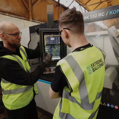 A male with a high vis vest and gloves explains digital readings on a machine screen to another person in a high vis jacket.