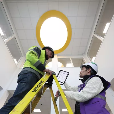 A male in a hard hat with a tablet shows the screen to another male in a hard hat standing on a ladder. 