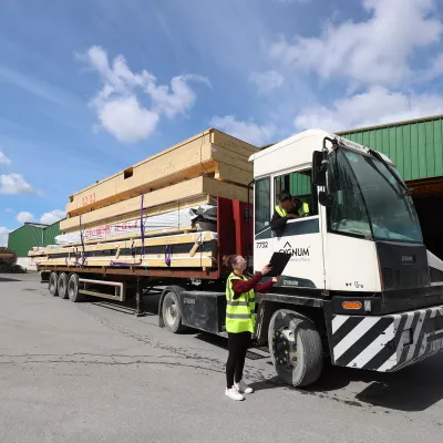 Person in high-vis gear standing beside a truck loaded with timber panels outside a warehouse.