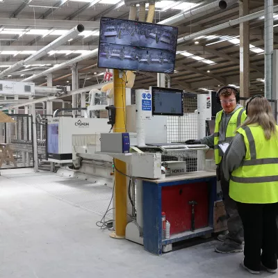 Two workers in high-vis vests monitoring a 3D printer control station inside a large manufacturing facility.