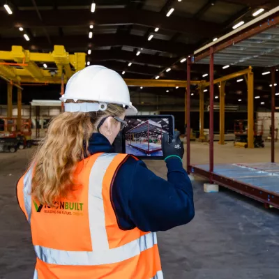 A female in a high vis jacket and white hard hat takes a picture using a tablet in a manufacturing facility.