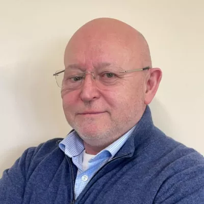 Headshot of a person wearing glasses and a blue sweater, standing in front of a plain light-coloured background.