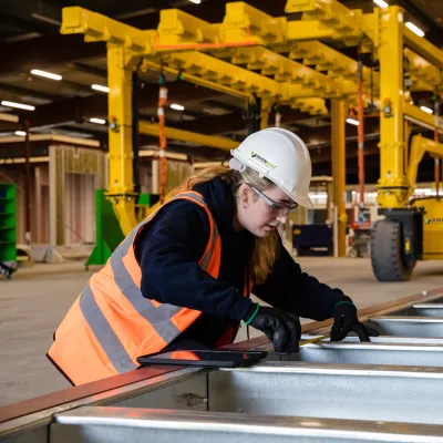A female in high vis vest and white hard hat measures a steel frame in a manufacturing facility.