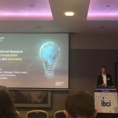 Person presenting at a conference next to a screen showing a slide with a lightbulb graphic.