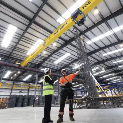 Two people wearing high vis jackets, hard hats and ear muffs inspect a crane in a manufacturing facility.
