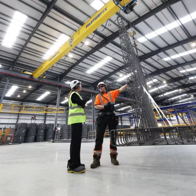 Two people in high-vis clothing standing inside a large factory, discussing equipment under an overhead crane.