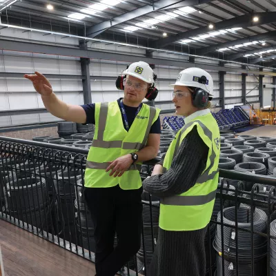 A male with a high vis vest, hard hat, and ear muffs points at something to a female in a high vis vest, hard hat, and ear muffs.