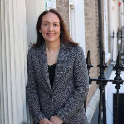Headshot of a person in a grey jacket standing outside a building.
