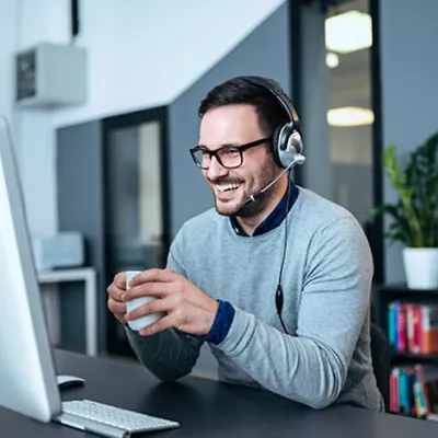 A male smiling holding a white mug sitting at a computer. He has black glasses and headphones.