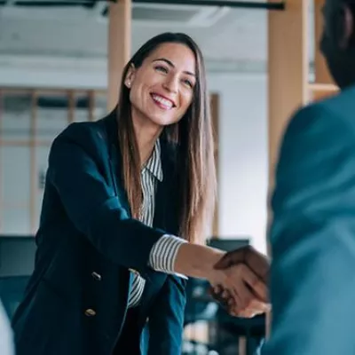 A female wearing a business suit smiling and shaking another person's hand.