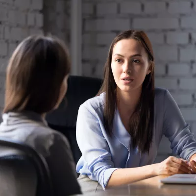 Two females in business clothing sit at a desk in a modern office space talking.