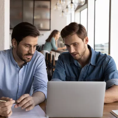 Two males in business shirts sit at a desk and read from a laptop. One takes notes with a pen on a piece of paper.