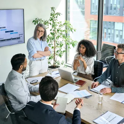 A group of people in business clothing with laptops and pieces of paper in front of them sit around a long table, talking.