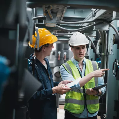 Two people in hard hats and work overalls, one holding a clipboard and the other holding a tablet, inspect machinery.