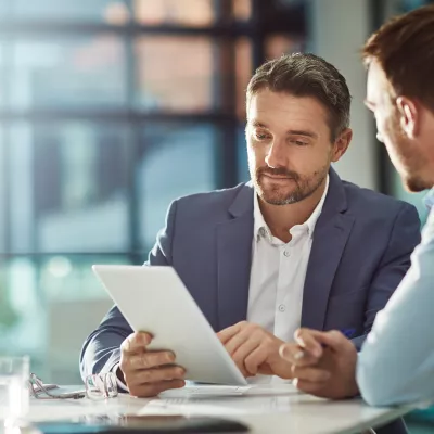 A male in a blue business suit reads from a tablet to another male in a business shirt.