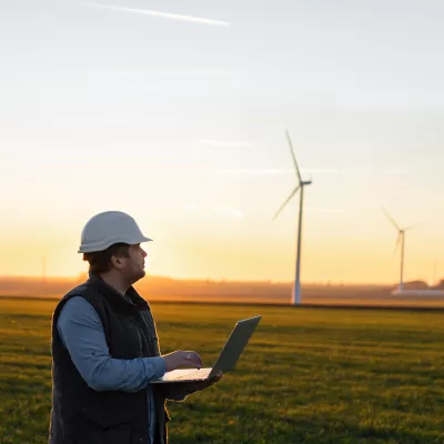 A male in hard hat and vest with an open laptop stands in a field with wind turbines in the background.