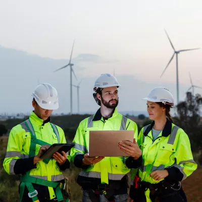 A group of people in high vis jackets and hard hats read from tablets standing in a field with wind turbines.