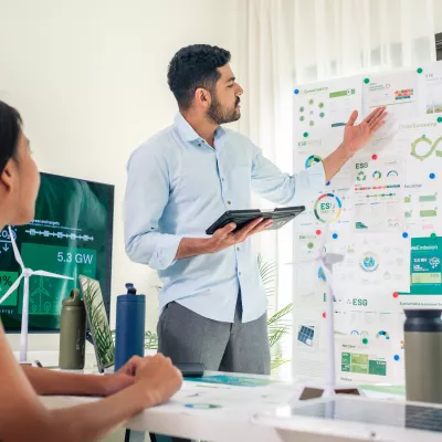 A male in a business shirt stands with a tablet in his hand explaining information on a flip chart to a group of people sat at a table. 