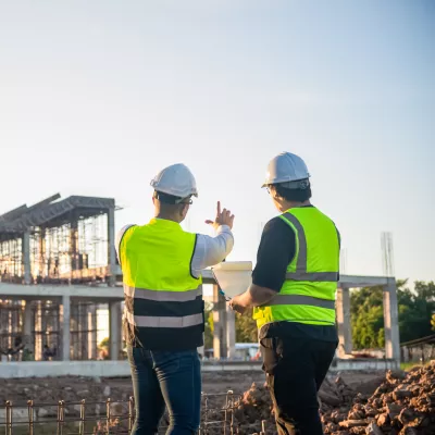 Two people in high-vis vests and hard hats standing at a construction site, looking at a building under development.