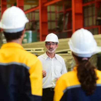 A male with glasses and a hard hat addresses a crowd of people wearing work overalls and hard hats.