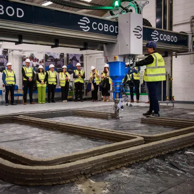 A group of people wearing high-visibility vests and hard hats standing in a row inside an industrial or construction facility, with machinery visible in the foreground.