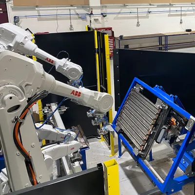 Industrial robotic arms working on components mounted in blue frames inside a manufacturing or training facility. Workbenches and tools are visible in the background.