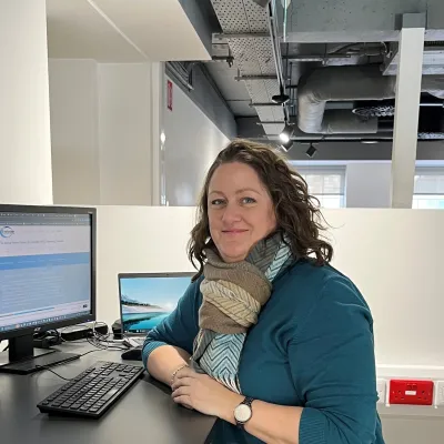 A person wearing a teal sweater and patterned scarf is seated at a desk, smiling at the camera, with two computer monitors in front of them in a modern office space.