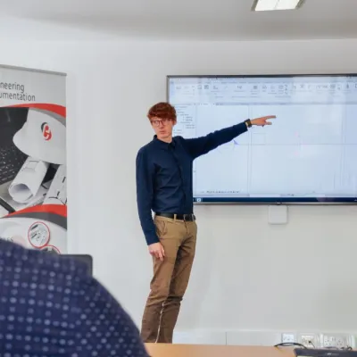 A person is standing next to a screen, pointing at a slide during a training session in a classroom.