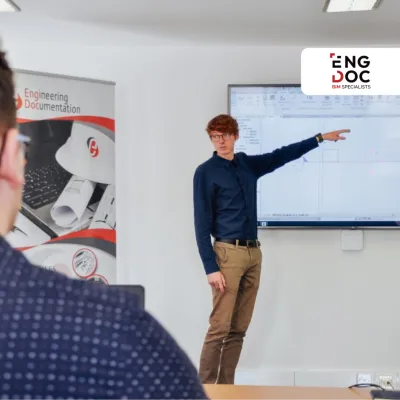 A person is standing next to a screen, pointing at a slide during a training session in a classroom.