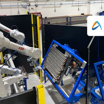Industrial robotic arms working on components mounted in blue frames inside a manufacturing or training facility. Workbenches and tools are visible in the background.