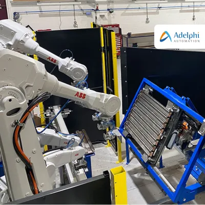 Industrial robotic arms working on components mounted in blue frames inside a manufacturing or training facility. Workbenches and tools are visible in the background.