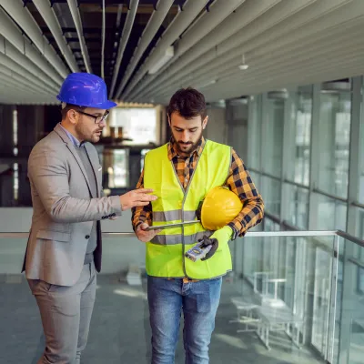 Two people wearing hard hats are standing on a construction site, discussing a project.