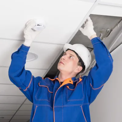 Worker in hard hat installing or inspecting a ceiling smoke detector.
