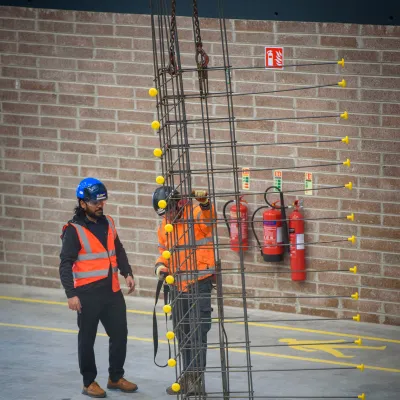Two construction workers in safety gear standing near a vertical rebar structure inside a building under construction.