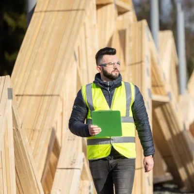 Construction worker wearing a high-visibility vest and holding a clipboard, standing in front of wooden roof trusses at an outdoor building site.