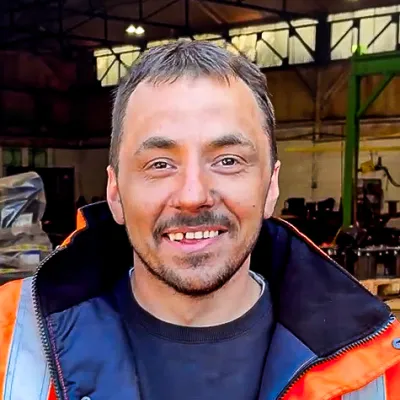 A person in a high-visibility jacket smiling indoors at an industrial or workshop setting.