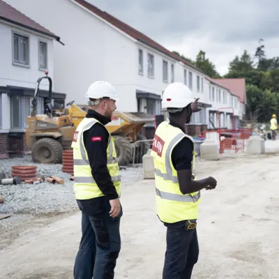 Three construction professionals in safety gear and hard hats are standing on a residential building site, discussing progress. Houses under construction are visible in the background.